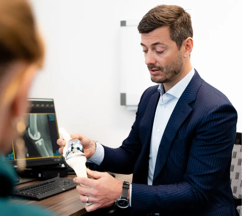 Orthopaedic specialist in navy pinstripe suit demonstrating knee joint model to patient during consultation