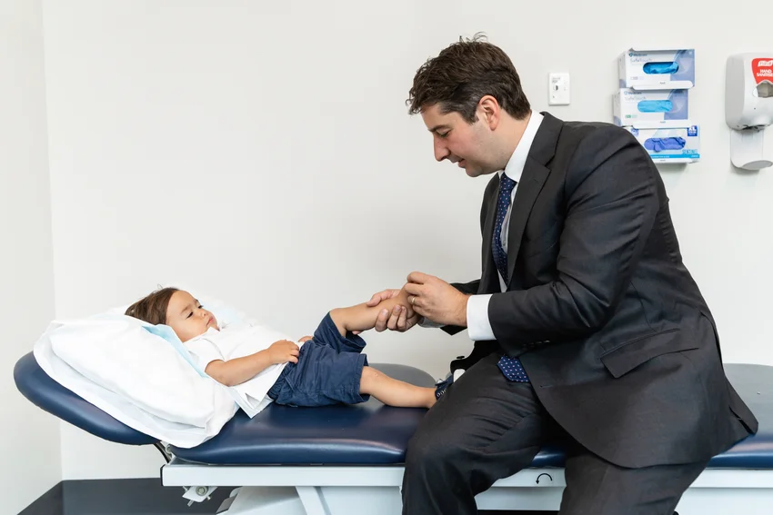 Paediatric orthopaedic specialist examining young patient's leg mobility during clinical assessment, with child resting on examination table in modern medical facility