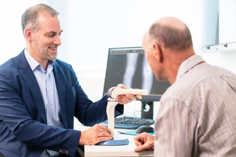 Orthopaedic surgeon demonstrating knee joint model to elderly patient during consultation with digital X-ray visible on computer monitor in modern clinical setting