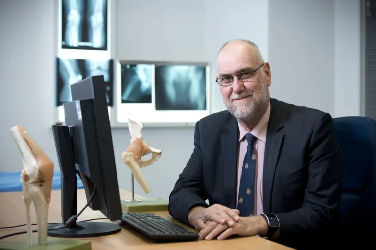 Orthopaedic surgeon seated at desk with anatomical spine model and diagnostic imaging displayed on wall behind