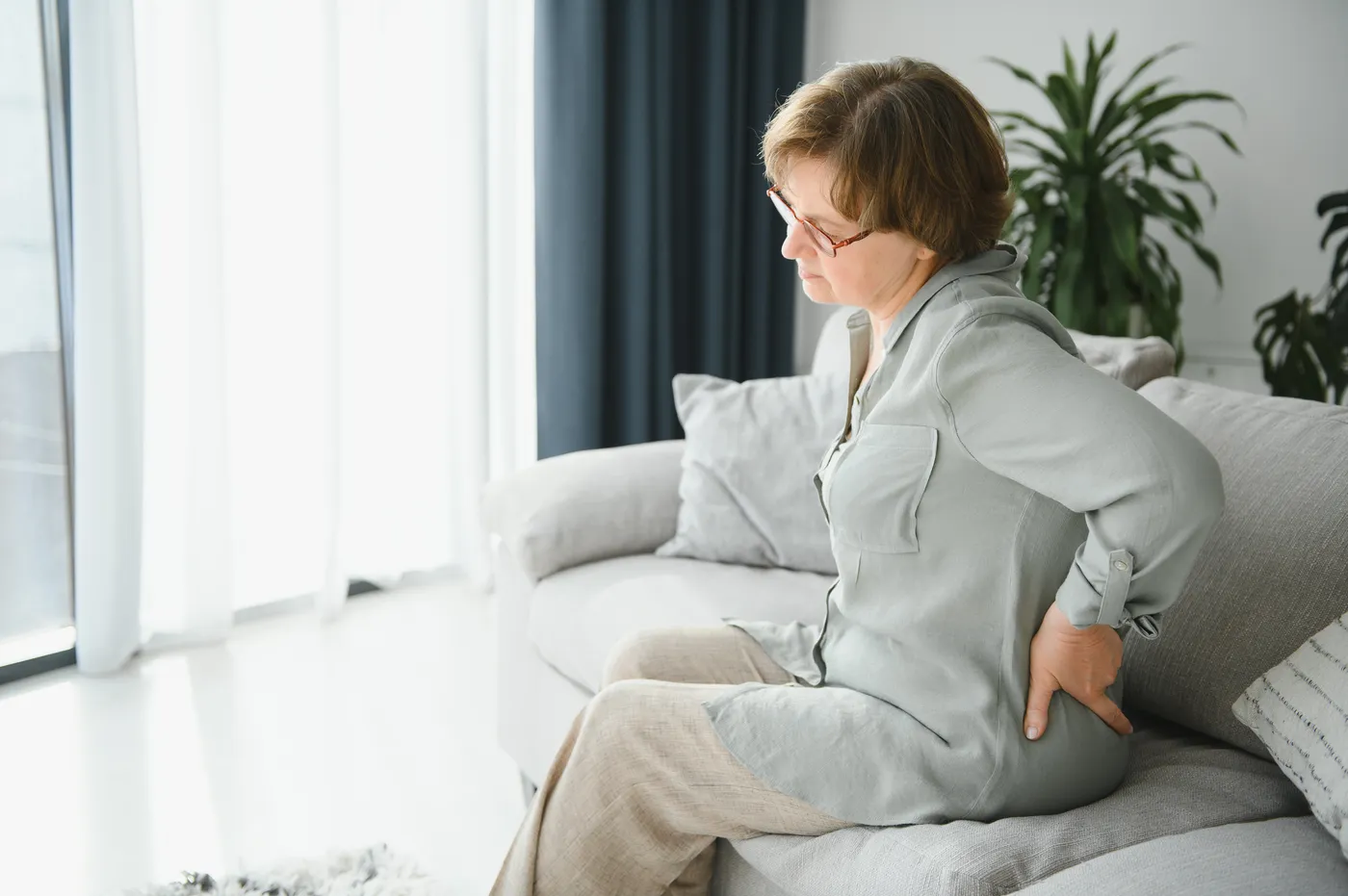 Woman with short brown hair and glasses experiencing back pain while sitting on couch in modern living room