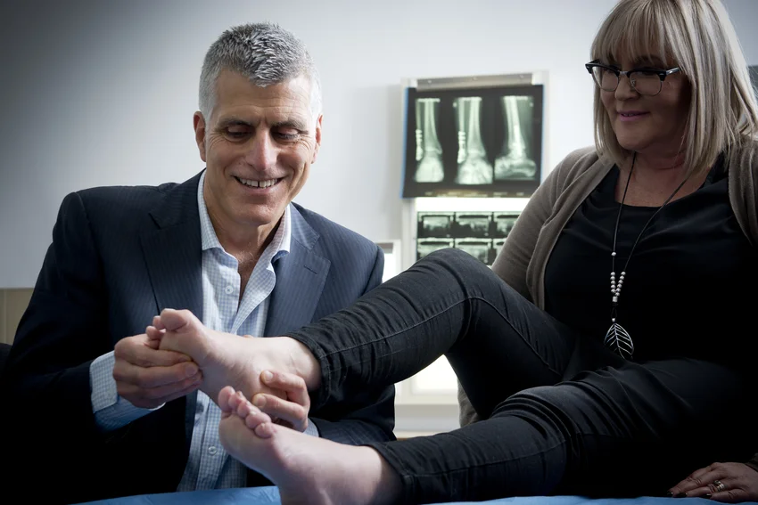 Podiatric specialist examining female patient's foot and ankle mobility during clinical assessment with foot X-rays displayed in background